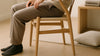 An adult casually sitting in a wishbone-style chair in a bright Scandinavian living room, highlighting comfortable posture and realistic wood and cord textures.